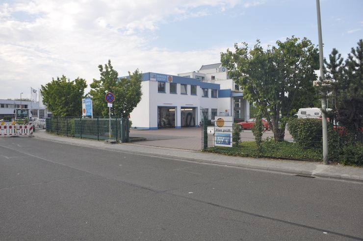 Ein weiß-blaues Gebäude mit dem Schild "TUEV Hessen", umgeben von grünen Bäumen und einem Zaun. Vor dem Gebäude ist ein rotes Auto geparkt.