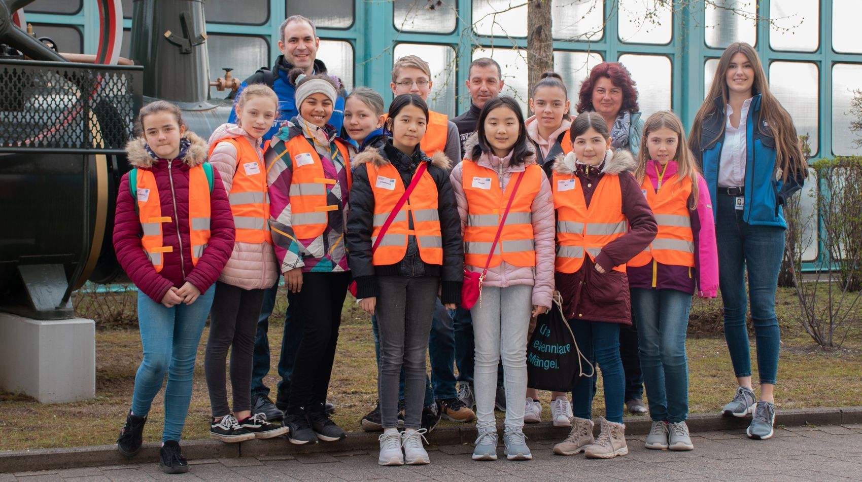 Eine Gruppe meist junger Menschen mit leuchtend orangefarbenen Sicherheitswesten steht vor einem Gebäude mit großen Fenstern.