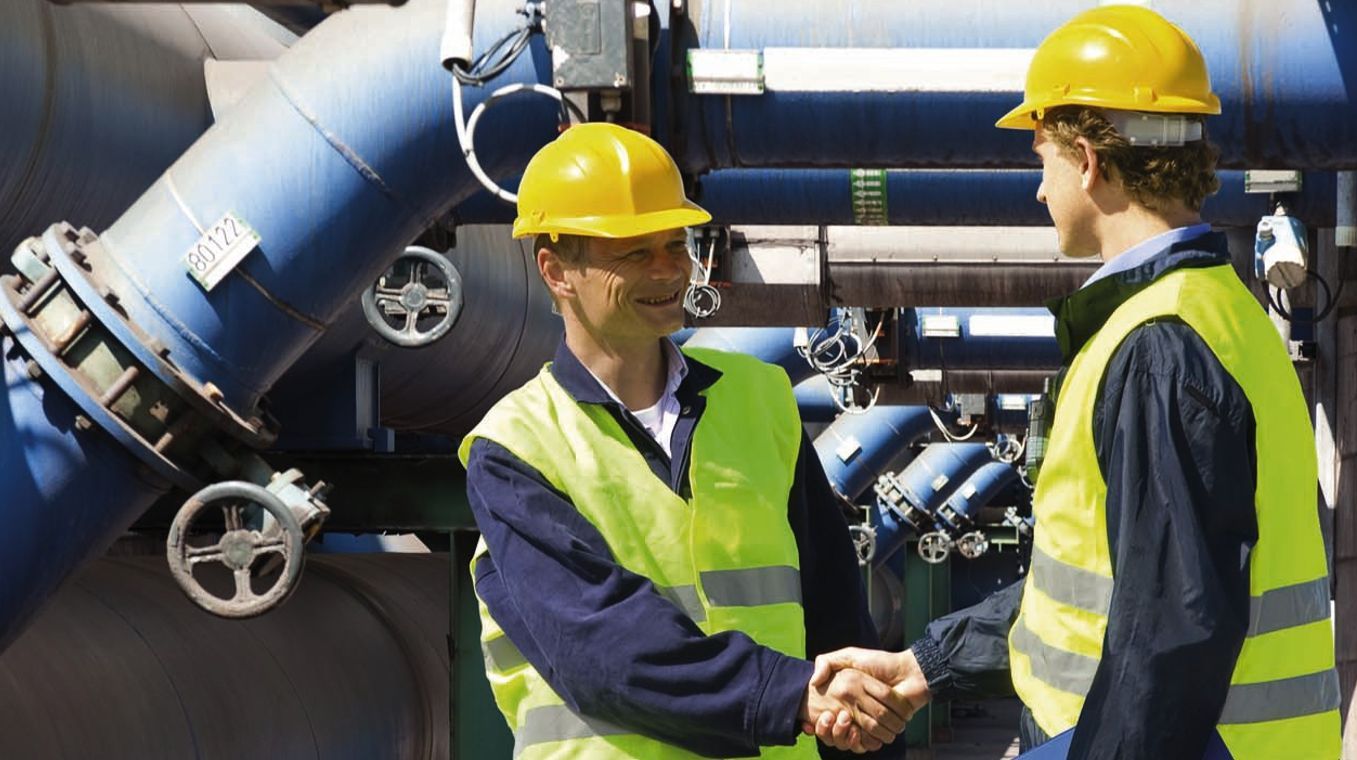 Zwei Männer in Sicherheitswesten und Schutzhelmen geben sich vor großen blauen Rohren die Hand.