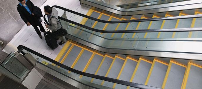 Zwei Personen auf einer Rolltreppe, von denen eine einen Koffer in der Hand hält, stehen auf einer grau-gelben Treppe.