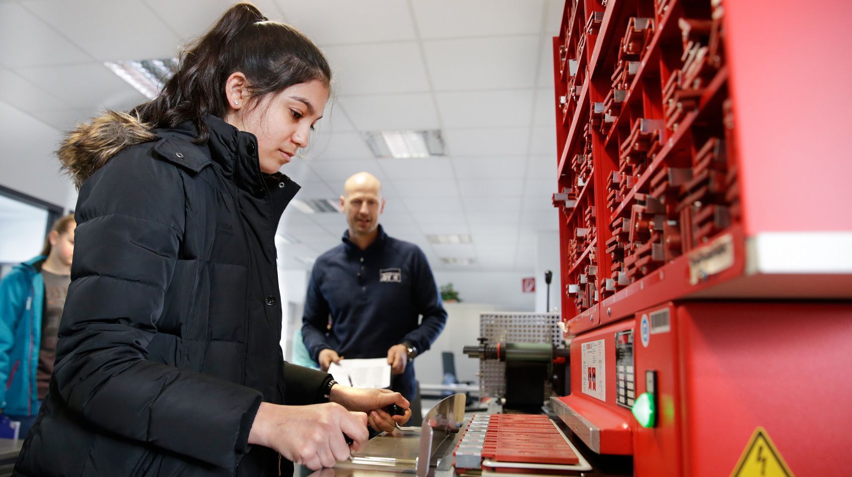 Eine junge Frau in einer schwarzen Jacke arbeitet an einer roten Maschine, ein Mann in einem blauen Hemd beobachtet sie dabei.