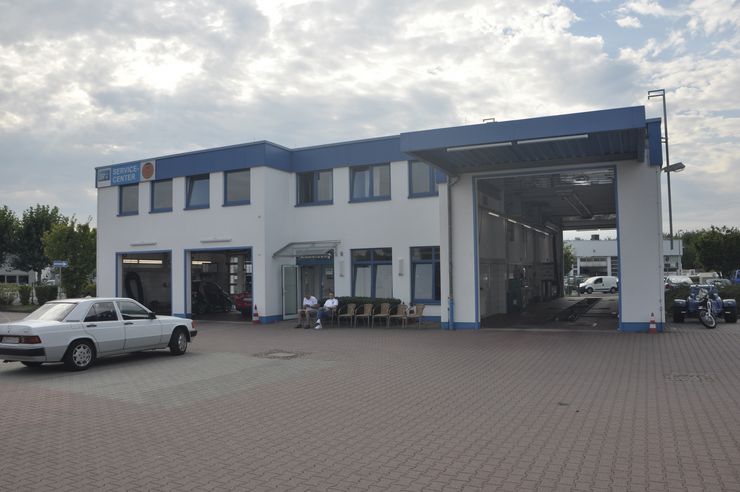 Ein weißes Gebäude mit blauer Umrandung und der Aufschrift "TUEV Hessen Service Center", das auf einem offenen Gelände steht, vor dem ein Auto geparkt ist. Vor dem Gebäude sitzen ein paar Personen.