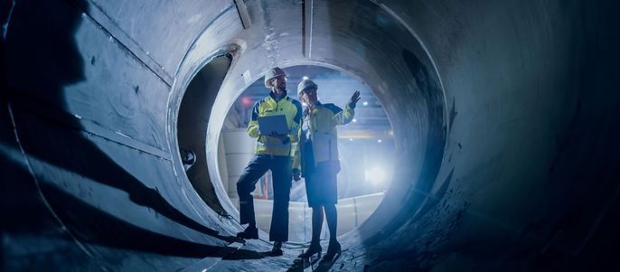 Zwei Personen, ein Männchen und ein Weibchen, stehen in einem großen zylindrischen Tunnel. Das Männchen deutet auf etwas, während das Weibchen es beobachtet.