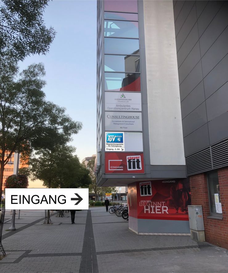Ein modernes Gebäude mit verschiedenen Werbungen und Schildern, darunter ein "Consultinghouse". Ein Richtungspfeil zeigt nach rechts auf den Eingang von einer TUEV Hessen Filiale, und man sieht eine Person auf dem Bürgersteig gehen.