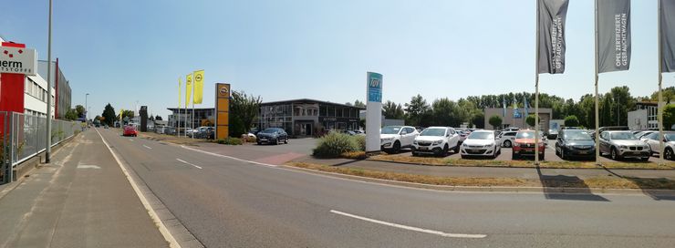 Ein Autohaus mit mehreren geparkten Autos, ein paar Fahnen und ein Gebäude mit dem Logo des TUEV Hessen.