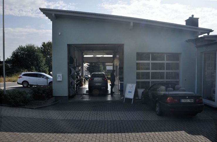 Eine Autowerkstatt mit einem hellblauen Gebäude, einem schwarzen Auto in der Garage, einem weißen Auto, das vor der Garage geparkt ist, und einem Schild vor der Garage.
