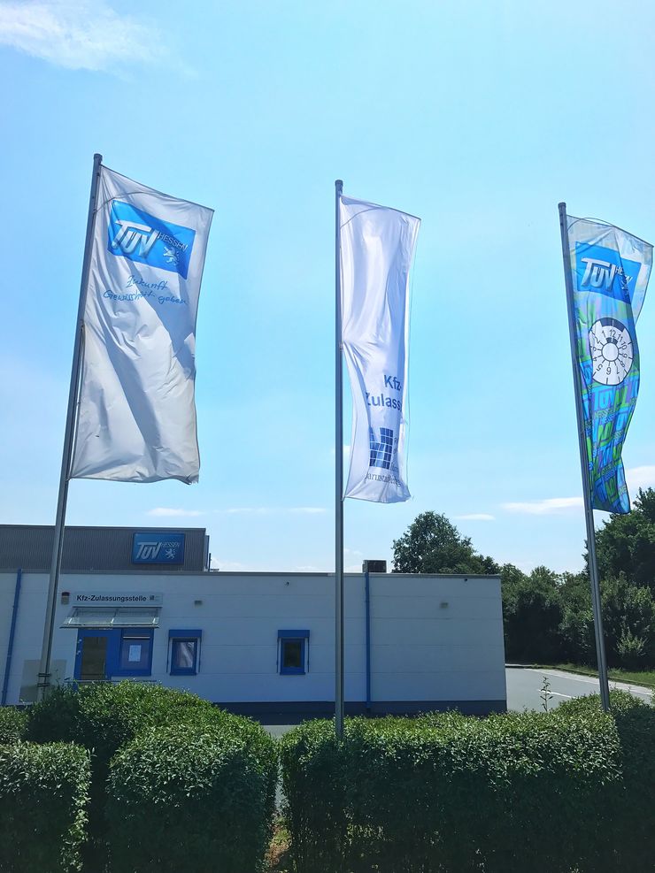 Drei Fahnen mit dem "TUEV Hessen"-Logo, ein Gebäude mit dem "TUEV Hessen"-Logo und ein Schild mit der Aufschrift "Kfz-Zulassungsstelle".