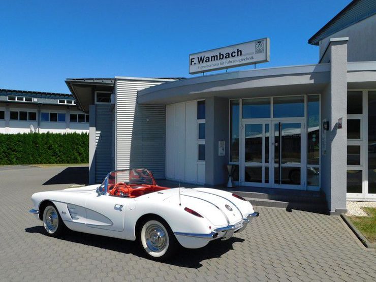 Ein klassisches weißes Auto parkt vor einem modernen Gebäude mit einem Schild, auf dem "F. Wambach' steht.