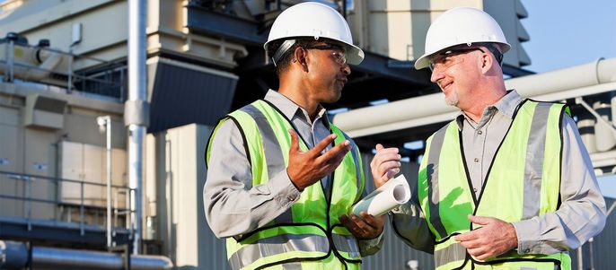 Zwei Männer, die Schutzhelme und Warnwesten tragen, unterhalten sich in einem Industriegebiet.