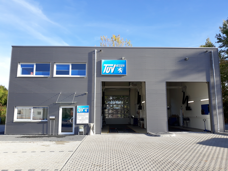 Ein modernes Gebäude mit dem TUEV Hessen-Logo an der Fassade in einer sonnigen Umgebung mit Bäumen im Hintergrund.