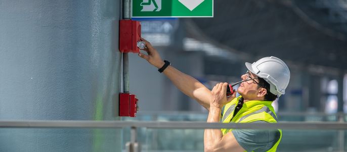 Ein Mann mit Sicherheitsweste und Helm, der einen roten Feuermelder bedient, der an einem grauen Mast befestigt ist.