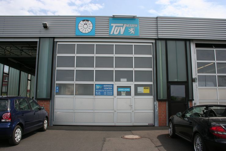 Ein Gebäude mit einem blauen Schild mit der Aufschrift "TUEV HESSEN" und dem Logo einer Uhr. Draußen sind zwei Autos geparkt, und das Gebäude hat ein Service-Center-Schild mit verschiedenen Details.