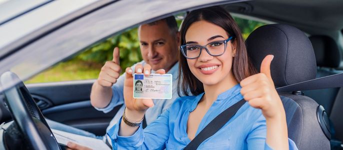 Eine Frau und ein Mann in einem Auto, beide zeigen den Daumen nach oben. Die Frau hält einen Führerschein mit einem Foto eines Mannes und einer Frau in der Hand.