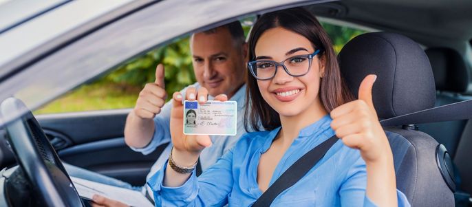Eine Frau und ein Mann in einem Auto, beide zeigen den Daumen nach oben. Die Frau hält einen Führerschein mit einem Foto eines Mannes und einer Frau in der Hand.