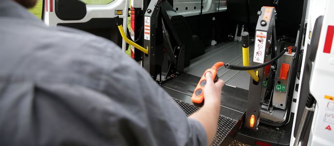 Eine Person, die ein Werkzeug in der Hand hält, steht neben einem Fahrzeug mit einer Rampe und verschiedenen Sicherheitsschildern.