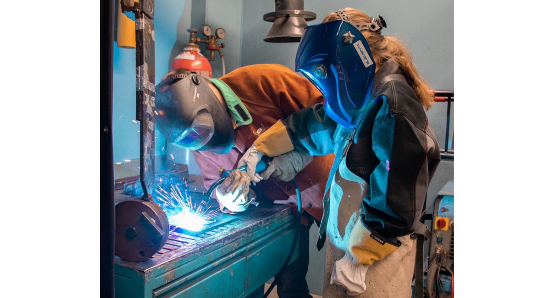 Zwei Personen in einer Werkstatt, die schweißen. Eine Person trägt einen Schutzhelm und orangefarbene Kleidung, während die andere einen blauen Helm und eine grüne Jacke trägt. Beide sind auf ihre Aufgabe konzentriert, während beim Schweißen Funken fliegen.