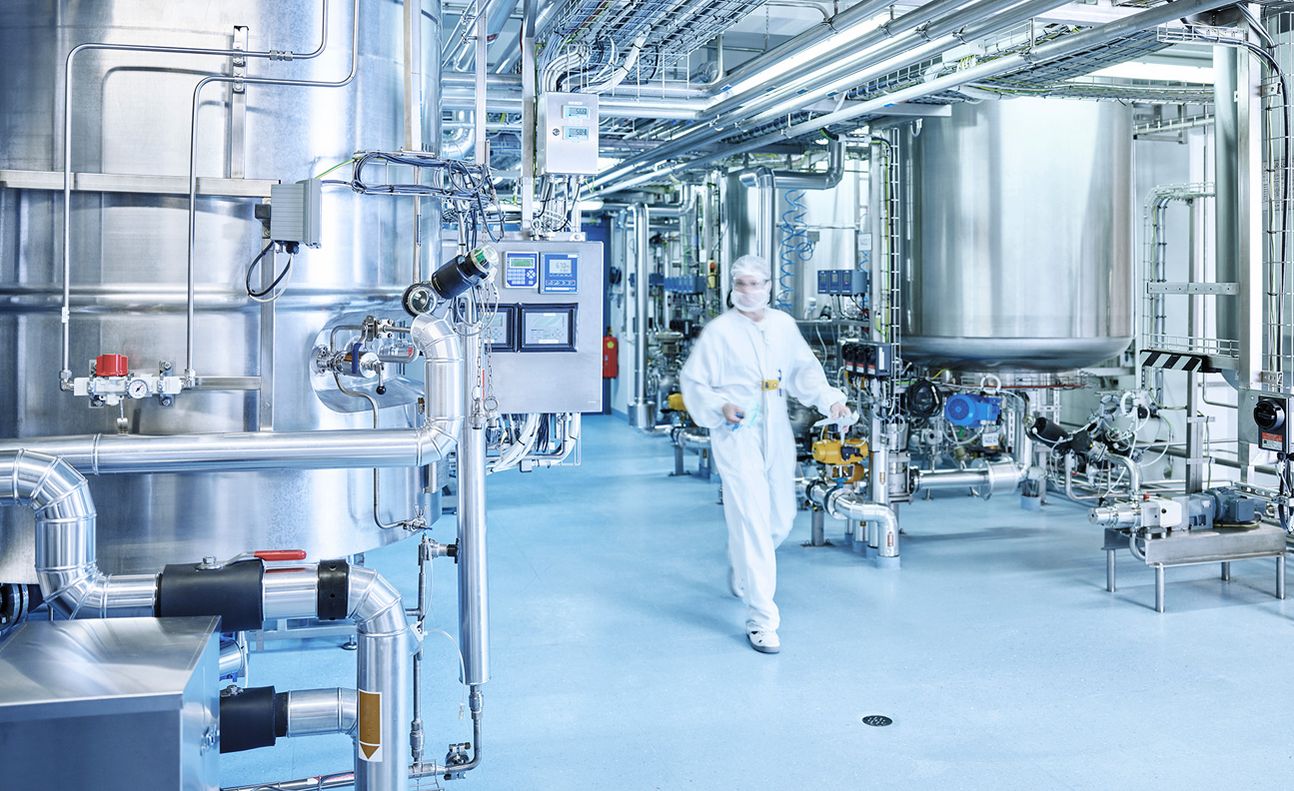 Eine Person in Schutzkleidung geht durch einen sauberen Industrieraum mit Metalltanks und Rohrleitungen.