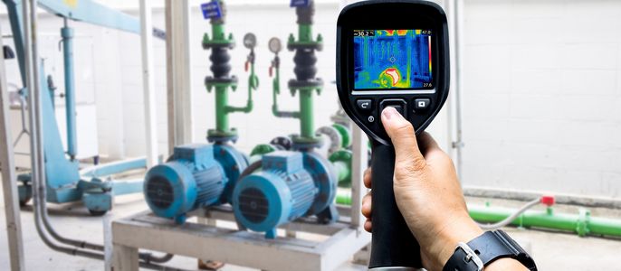Eine Hand, die ein Wärmebildgerät hält und eine Ansicht eines mechanischen Systems mit blauen Pumpen und grünen Rohren aufnimmt.