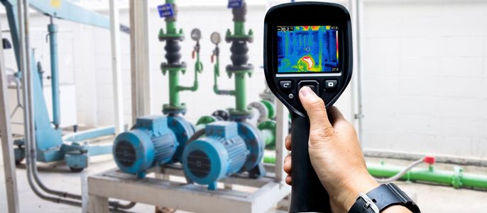 Eine Hand, die ein Wärmebildgerät hält und eine Ansicht eines mechanischen Systems mit blauen Pumpen und grünen Rohren aufnimmt.