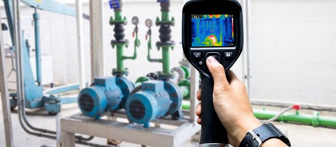 Eine Hand, die ein Wärmebildgerät hält und eine Ansicht eines mechanischen Systems mit blauen Pumpen und grünen Rohren aufnimmt.