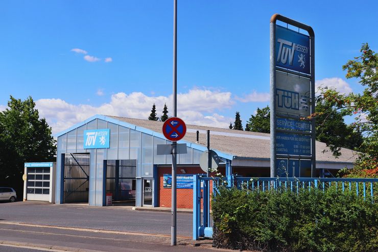 Eine Autowaschanlage namens "TUEV Hessen". Draußen parkt ein blaues Auto, und die Umgebung besteht aus Bäumen und einem klaren blauen Himmel.