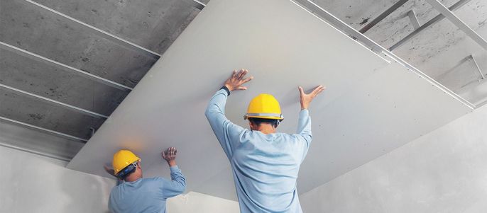 Zwei Arbeiter, die gelbe Helme tragen, montieren eine weiße Deckenplatte in einem Raum mit Sichtbetonbalken.