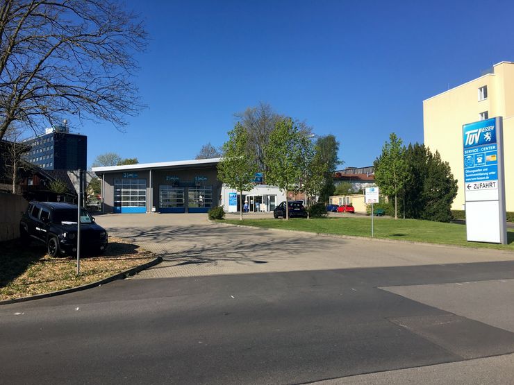 Eine moderne Tankstelle mit einem blauen Schild, ein Gebäude mit einem "TUEV"-Logo und ein paar geparkte Autos davor.