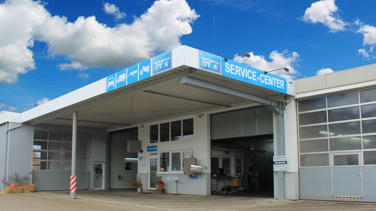 Ein modernes Gebäude mit einer blauen Markise mit der Aufschrift "TUEV Service-Center". Das Gebäude hat eine weiße Fassade, große Fenster und eine Tür mit der Aufschrift "ENGINE". Im Inneren des Gebäudes befinden sich ebenfalls einige Gegenstände und Geräte, und der Himmel über dem Gebäude ist teilweise bewölkt.