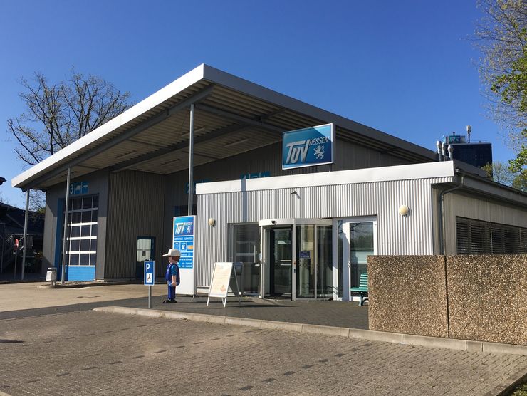 Ein modernes Gebäude mit einem blauen Schild mit der Aufschrift "TUEV" und dem Logo eines Autos. Die Umgebung besteht aus einem Parkplatz und Bäumen.