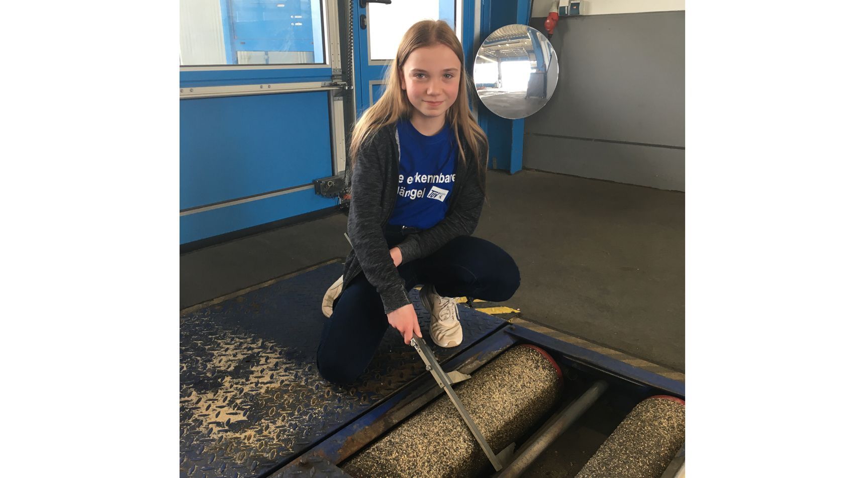 Eine junge Frau im blauen Hemd kniet auf einem blauen Podest und hält ein langes Werkzeug in der Hand, während ein Spiegel die Umgebung hinter ihr reflektiert.
