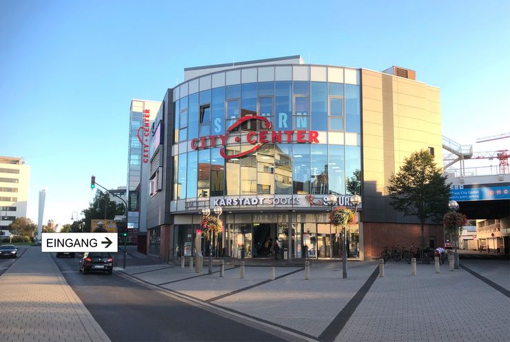 Ein modernes Gebäude mit der Aufschrift "City Center" und einem Schild mit der Aufschrift "Sport & City Center". Im Inneren befinden sich verschiedene Geschäfte und Einrichtungen, und ein Schild weist auf "Karstadt Sports" hin. Das Gebäude ist von Straßen und Fußgängerwegen umgeben, auf denen ein paar Leute herumlaufen.