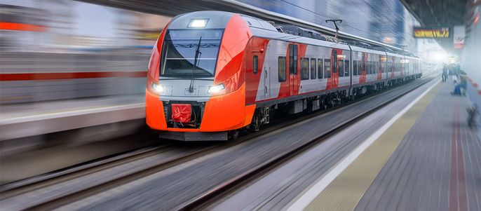 Ein schnell fahrender Zug mit leuchtend roter Front, der auf Gleisen fährt, mit einem unscharfen Hintergrund aus einem Bahnhof und Gebäuden.