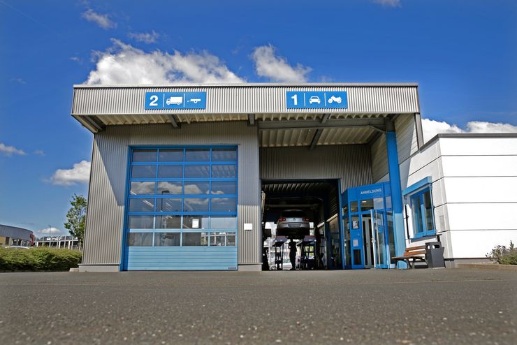 Ein modernes Gebäude mit zwei Garagentoren, eines mit der Aufschrift '2' und das andere mit der Aufschrift '1', unter einem blauen Dach. Auf der rechten Seite befindet sich ein Schild mit der Aufschrift "ANMELDUNG".