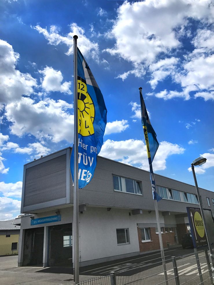 Zwei blaue Fahnen mit gelben Abbildungen wehen vor einem modernen Gebäude mit der Aufschrift "Hier prueft TUEV Hessen".