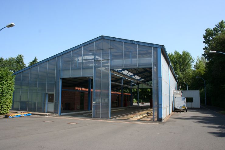 Ein großes Industriegebäude mit einem transparenten Dach, das auf einer Freifläche liegt, umgeben von Bäumen und einer Straße.