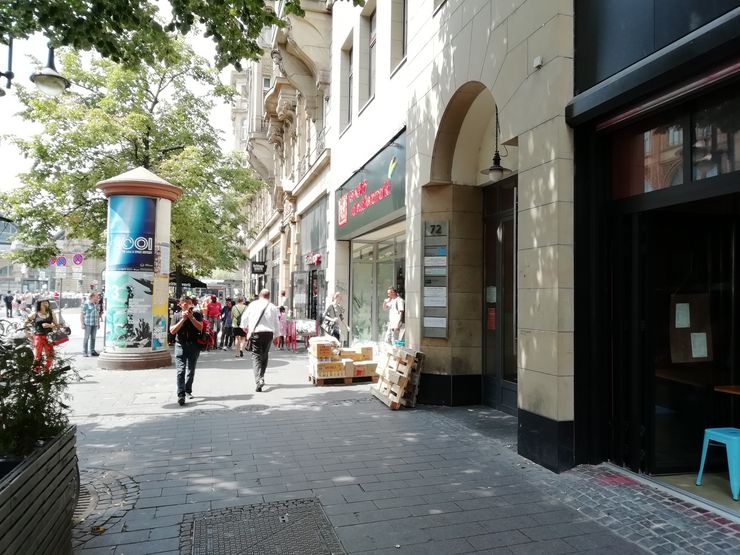 Eine belebte städtische Straße mit Menschen, die spazieren gehen, eine Schaufensterfront mit einem Schild und ein zylindrisches Gebäude mit einer Werbung.