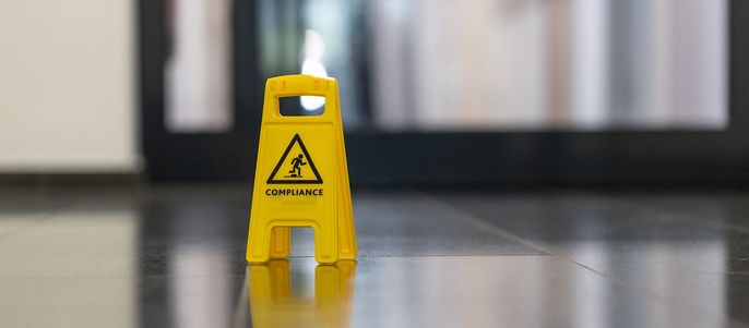 Ein gelbes "COMPLIANCE"-Schild auf einem glänzenden Boden, mit einem unscharfen Hintergrund eines Gebäudes.