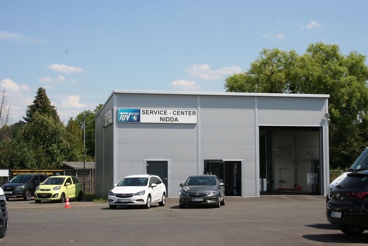 Ein Gebäude mit der Aufschrift "TUEV Hessen Service Center Nidda" und einem Schild darüber. Vor dem Gebäude sind mehrere Autos geparkt, darunter ein leuchtend gelbes, ein weißes und ein schwarzes.