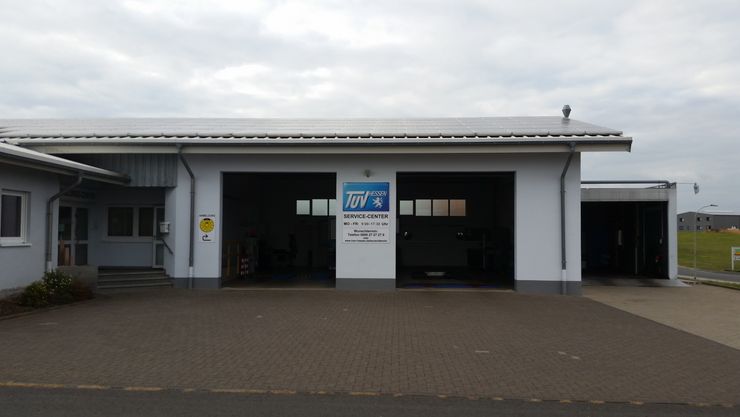 Ein Gebäude mit einem blauen Schild mit der Aufschrift "TUEV Hessen Service Center" und einer Telefonnummer. Das Gebäude hat eine weiße Fassade und eine gemauerte Einfahrt.