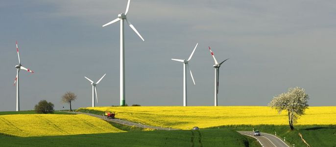 Eine malerische Landschaft mit einer Straße, die durch ein grünes Feld führt, flankiert von einem leuchtend gelben Feld und mehreren Windturbinen, die sich vor einem klaren blauen Himmel erheben.