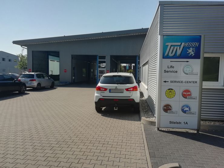 Ein Auto-Service-Center mit dem Namen 'TUEV HESSEN' mit einem Schild, das verschiedene Dienstleistungen wie 'Life Service', 'Service-Center' und 'Stielstr. 1A'. Draußen sind Autos geparkt, und das Gebäude hat ein modernes Design mit einer grauen Fassade.