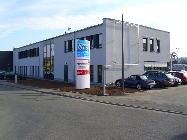 Ein modernes Gebäude mit einem Schild, auf dem "TUEV Hessen Service Center" steht. Das Gebäude hat mehrere Fenster und ist von geparkten Autos umgeben.