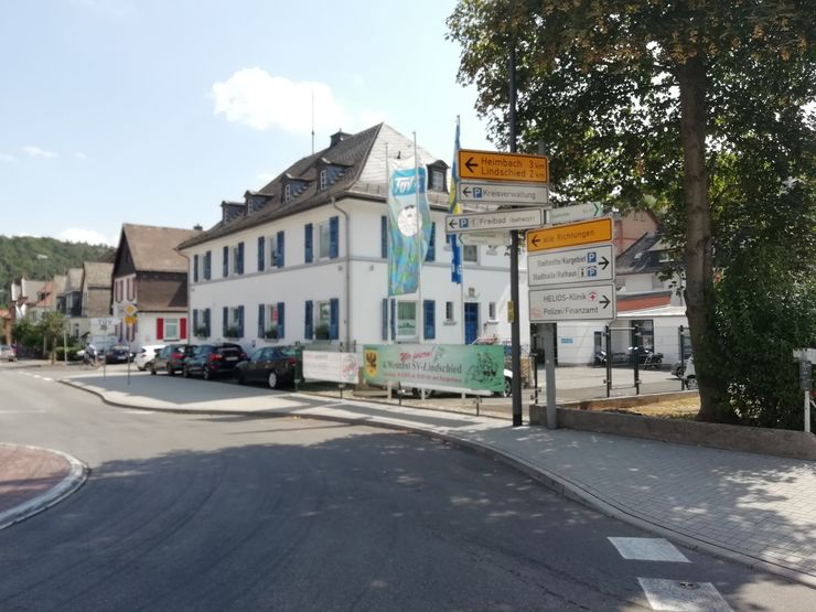Eine Straßenszene mit einem weißen Gebäude, einem Schild, geparkten Autos und einem Werbebanner für eine lokale Veranstaltung.