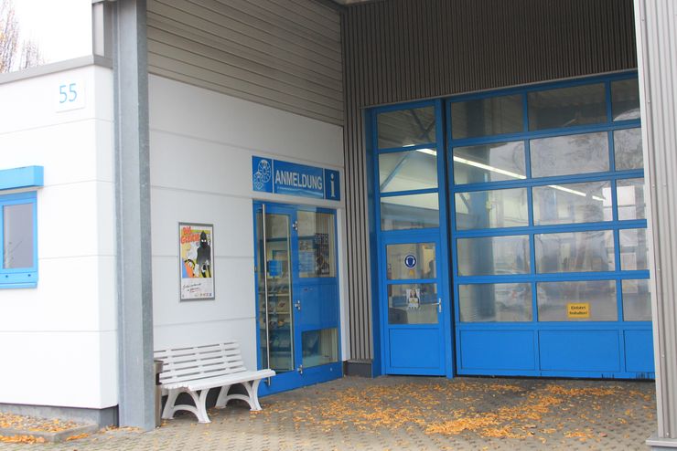 Ein weißes Gebäude mit einer blauen Tür mit der Aufschrift "ANMELDUNG" und einem Schild mit dem Bild einer Person. Vor dem Gebäude steht eine Bank, und der Boden ist mit gelben Blättern bedeckt.