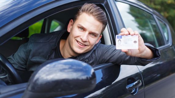 Junger Mann zeigt seinen Führerschein aus einem Auto lehnend.
