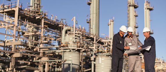 Eine Gruppe von Arbeitern auf einem Industriegelände, die einen Bauplan begutachten. Die Baustelle besteht aus hohen Strukturen, Rohren und Geräten. Die Arbeiter tragen Schutzhelme und Schutzkleidung.