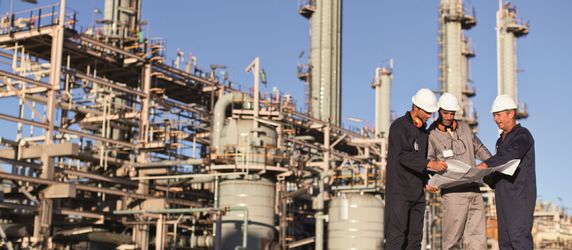 Eine Gruppe von Arbeitern auf einem Industriegelände, die einen Bauplan begutachten. Die Baustelle besteht aus hohen Strukturen, Rohren und Geräten. Die Arbeiter tragen Schutzhelme und Schutzkleidung.