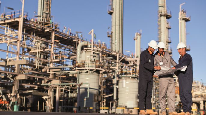 Eine Gruppe von Arbeitern auf einem Industriegelände, die einen Bauplan begutachten. Die Baustelle besteht aus hohen Strukturen, Rohren und Geräten. Die Arbeiter tragen Schutzhelme und Schutzkleidung.
