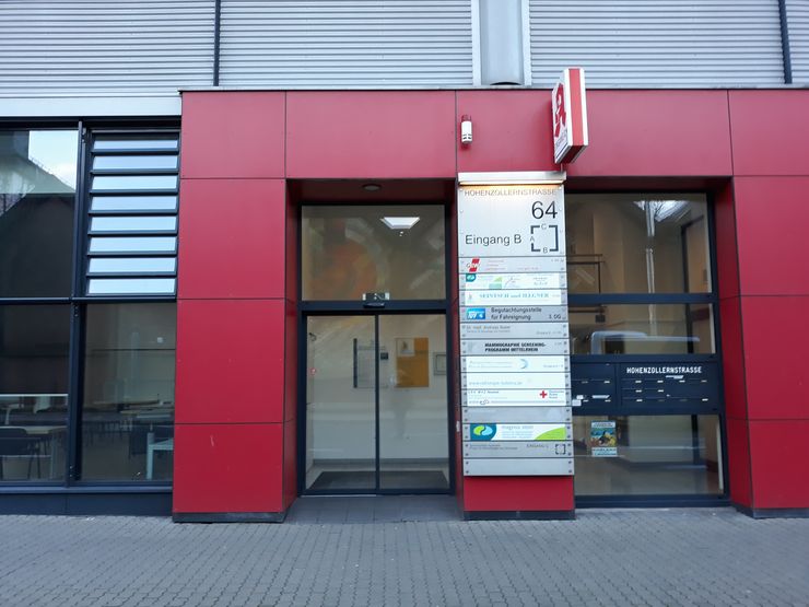 Ein modernes Gebäude mit roter Fassade, einem gläsernen Eingang und einem Schild mit verschiedenen Informationen und Wegbeschreibungen.