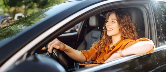 Eine Frau mit lockigem Haar, die ein orangefarbenes Oberteil trägt und ein schwarzes Auto fährt. Sie lächelt und schaut aus dem Fenster.